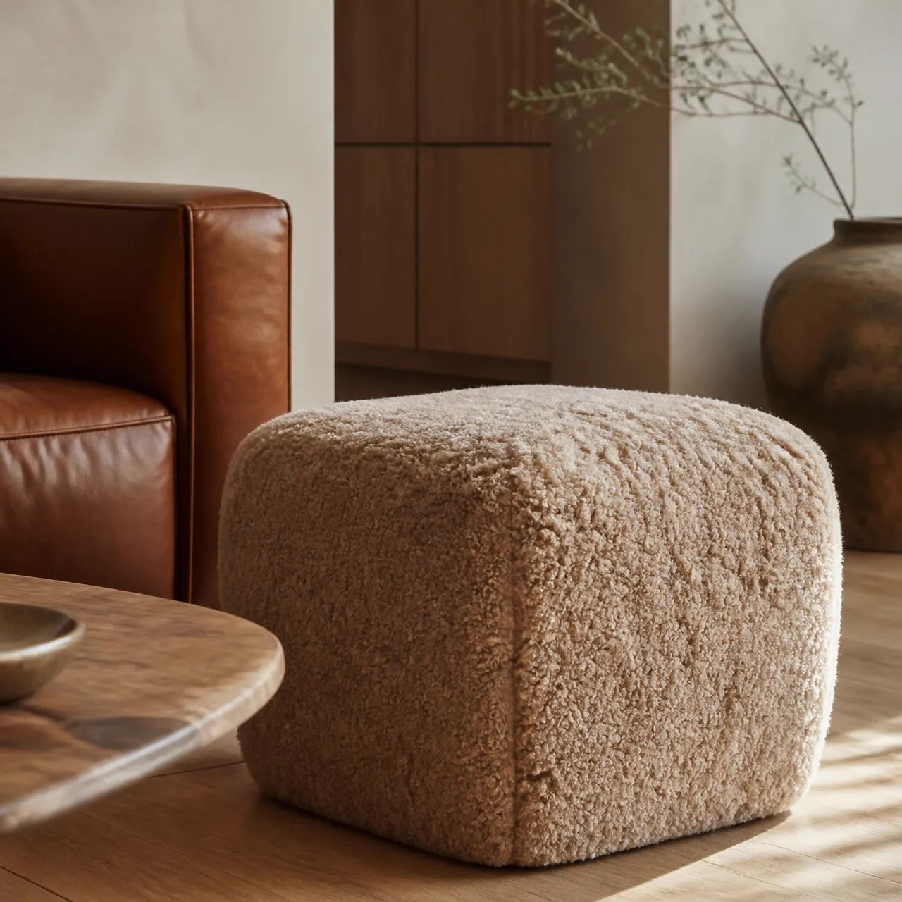 Sand-colored shearling pouf ottoman beside cognac leather seating in a warm residential interior